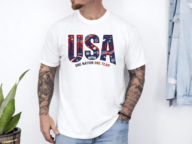 May include: A white short-sleeved t-shirt featuring the large blue and red text USA on the chest. The letters contain graphic illustrations of American landmarks, flags, and symbols. Below the text, the words ONE NATION ONE TEAM are printed.