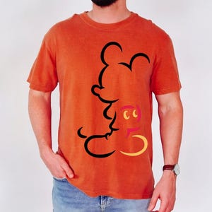 May include: Orange t-shirt featuring a black outline of Mickey Mouse. The shirt has a crew neck and short sleeves. The design includes yellow, pink, and black accents. The shirt is made of a soft material.