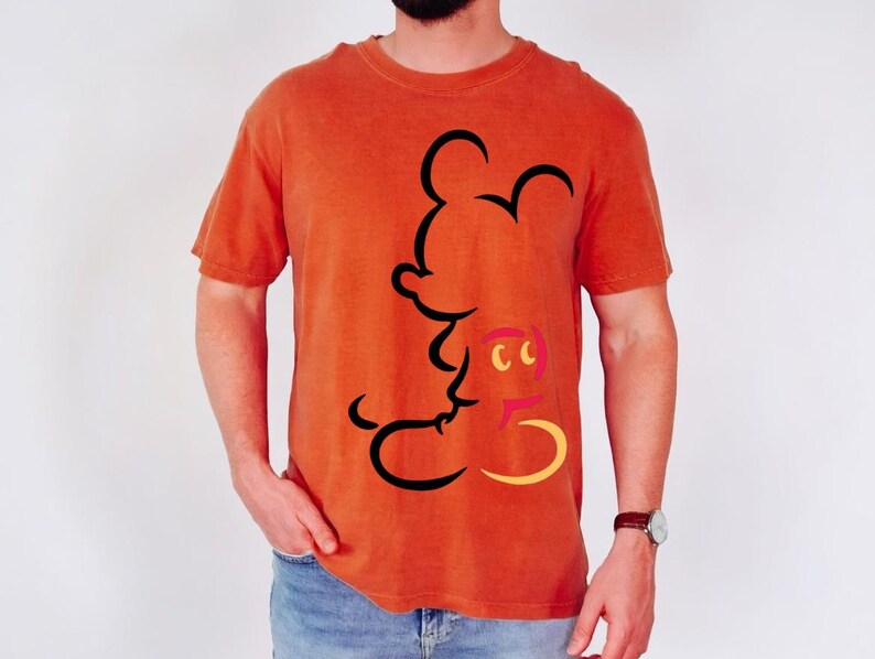 May include: Orange t-shirt featuring a black outline of Mickey Mouse. The shirt has a crew neck and short sleeves. The design includes yellow, pink, and black accents. The shirt is made of a soft material.