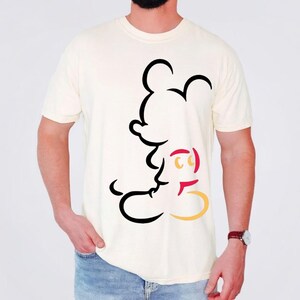 May include: Cream-colored t-shirt featuring a black, red, and yellow outline of Mickey Mouse. The graphic is on the front of the shirt. The shirt is being worn by a person in blue jeans.