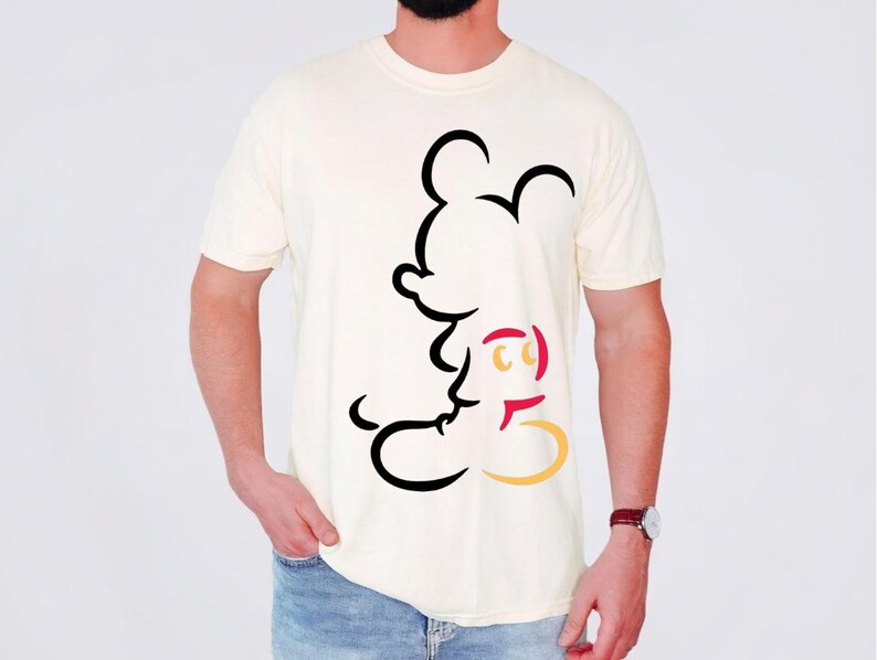 May include: Cream-colored t-shirt featuring a black, red, and yellow outline of Mickey Mouse. The graphic is on the front of the shirt. The shirt is being worn by a person in blue jeans.