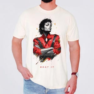 May include: A cream-colored short-sleeve t-shirt featuring a graphic print of a person with dark hair wearing a red jacket with black accents. The person has their arms crossed. Below the graphic, the text BEAT IT is printed in red capital letters.