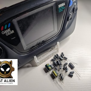 May include: A black Sega Game Gear portable video game system with a clear screen, a white stand, and a pile of capacitors. The Game Gear has the text "GAME GEAR" and "SEGA PORTABLE VIDEO GAME SYSTEM". A logo for "CAT ALIEN PRODUCTIONS" is in the lower left.