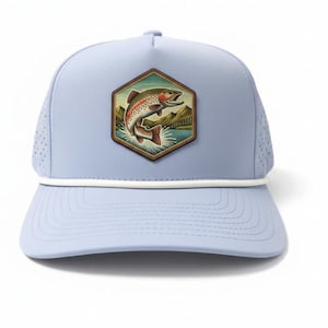 May include: A light blue baseball cap featuring a hexagonal patch on the front. The patch displays a colorful illustration of a jumping trout fish against a backdrop of mountains and water. A thin white cord runs across the base of the crown.