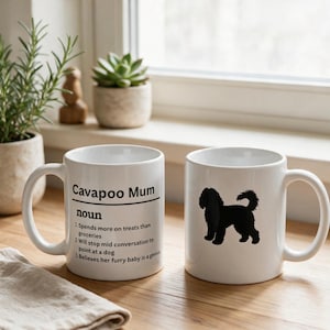 May include: Two white ceramic mugs. One mug features the black text 'Cavapoo Mum noun 1. Spends more on treats than groceries 2. Will stop mid conversation to point at a dog 3. Believes her furry baby is a genius'. The other mug shows a black silhouette of a dog.