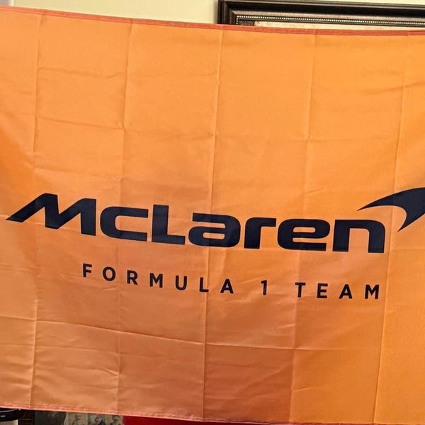 Puede incluir: Bandera naranja con el logotipo de McLaren Formula 1 Team en negro y azul marino. La bandera rectangular muestra el nombre del equipo y el texto « FORMULA 1 TEAM ». Fabricada en tela ligera.