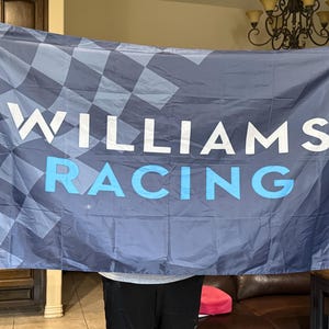 May include: A rectangular flag with the words "WILLIAMS RACING" in white and light blue. The flag has a checkered pattern in the upper left corner and a dark blue background.