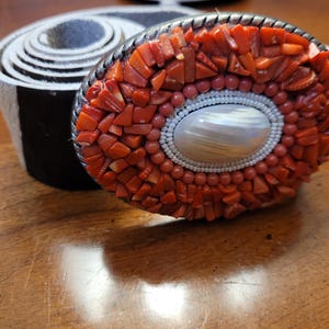 Peut inclure: Boucle de ceinture ovale ornée d'une mosaïque de morceaux de corail rouge, d'un ovale central en nacre et d'une bordure perlée. La boucle est sertie dans un cadre en métal argenté. Une ceinture rayée noire et blanche roulée est à côté.