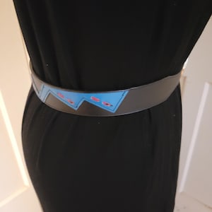 May include: A black leather belt with a blue and pink geometric design. The belt has a buckle and is worn around the waist.