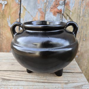 May include: A black ceramic cauldron with two handles and three short legs. The cauldron is sitting on a wooden surface.