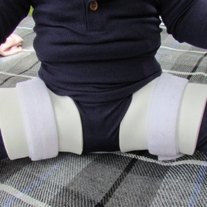 May include: A white and beige hip abduction brace with adjustable Velcro straps is worn by a child. The brace is designed to keep the legs in a specific position. The child is wearing a navy blue long-sleeved shirt and pants.