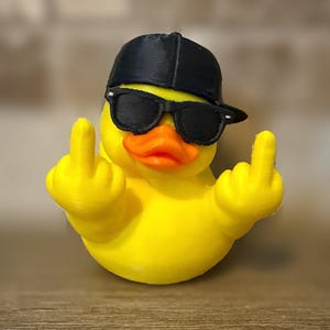 Op de afbeelding: Een felgeel rubberen eendje met een zwarte honkbalpet en een zwarte zonnebril. Het eendje heeft een oranje snavel en beide vleugels zijn omhoog geheven, waarbij elke vleugel een opgestoken middelvinger naar de kijker toe laat zien.