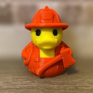 May include: A yellow rubber duck wearing an orange firefighter helmet and uniform, including an axe. The duck has black eyes and a yellow beak. The firefighter duck is a novelty item.
