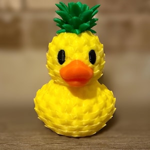 Op de afbeelding: Een gele eendenfiguur met een groene ananas bovenop. De eend heeft zwarte ogen en een oranje snavel. De ananas bovenop is groen en getextureerd. Het lichaam van de eend is ook getextureerd, wat het een unieke uitstraling geeft.