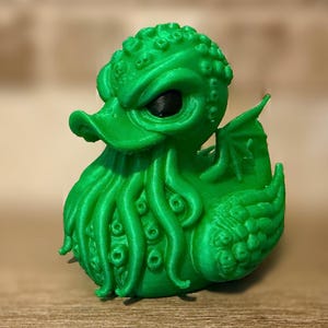 Puede incluir: Una figura de pato de color verde brillante impresa en 3D con una cabeza texturizada con protuberancias, ojos negros y un pico rodeado por múltiples tentáculos colgantes. El pato tiene pequeñas alas en los costados y un cuerpo texturizado.