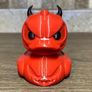 Puede incluir: Un pato de goma rojo con cuernos negros y ojos enojados. El pato es un juguete con temática de Halloween.