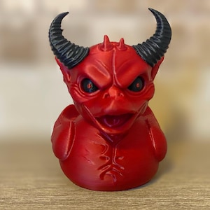 May include: A red devil duck figurine with black horns and angry eyes. The duck has a detailed, textured surface and a menacing expression. A unique and quirky decorative item.