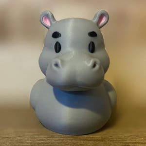 May include: A gray hippo-shaped figurine with black eyes and pink-lined ears. The hippo has a rounded body and a smiling expression. The figurine is sitting on a wooden surface.