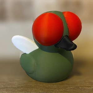 May include: A 3D-printed fly figurine with large, red eyes and a black beak. The body is green, with a white wing on one side. The figurine is designed to resemble a cartoon fly.