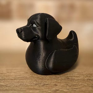 May include: A black Labrador Retriever duck figurine. The duck-shaped figurine has a detailed head, floppy ears, and a rounded body. The dog duck is made of a solid black material and sits on a wooden surface.