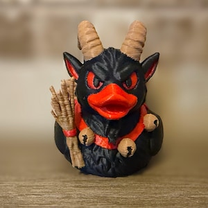 May include: A black rubber duck figurine featuring tan horns, a bright red beak, and red eyes. The duck wears a red collar with small bells and holds a tan broom in its wing. The surface has a textured, 3D-printed appearance with visible layers.