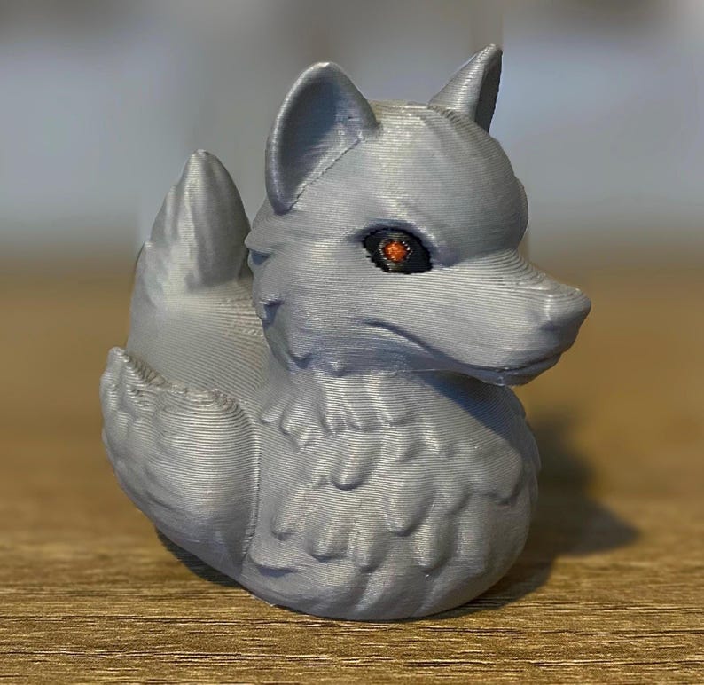 A gray 3D printed duckies shaped like wolves with red eyes. The duckies are sitting on a wooden surface. Rubber Duck style. Facing right.