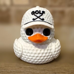May include: A white rubber duck with a dimpled golf ball texture, featuring a bright orange beak and black eyes. The duck wears a matching white golf cap with the word GOLF embroidered in black above two crossed golf clubs on the front panel.