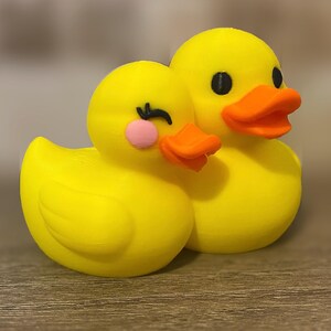 3D printed couples Duck of a girlfriend and a boyfriend all yellows ducks connected with orange beaks and black eyes. Girlfriend duck has eyelashes and pink Rosie cheek. Rubber Duck style perfect for jeep ducking. Facing right front.