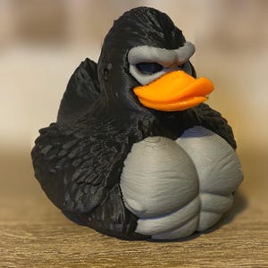 Könnte beinhalten: Eine neuartige Gummiente mit einem gorillaähnlichen Design. Die Ente hat einen schwarzen Körper, eine graue Brust und Augenbrauen sowie einen orangefarbenen Schnabel. Dies ist ein einzigartiger und humorvoller Dekorationsartikel.