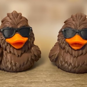 May include: Two brown duck figurines with textured feathers, bright orange beaks, and black sunglasses. The duck on the left is making a hand gesture with its wing, while the duck on the right has both wings tucked at its sides.