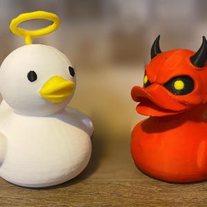 May include: Two novelty duck figurines: one white with a yellow halo and beak, the other red with black horns and yellow eyes. The angel duck and devil duck are side-by-side.