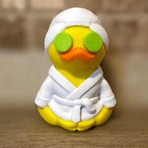 以下が含まれることがあります： 白いバスローブ、白いヘッドラップ、緑のキュウリのスライスを目の上に着けた黄色のカモの置物。カモはオレンジ色のくちばしをしています。スパをテーマにしたアイテムです。