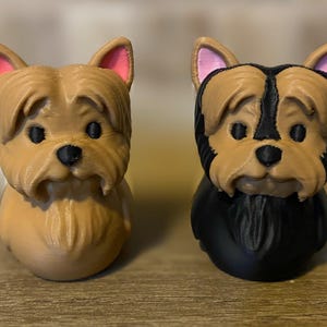 Może przedstawiać: Dwie figurki psów wydrukowane w 3D. Jedna jest w kolorze brązowym z różowymi uszami, druga jest czarno-brązowa z różowymi uszami. Obydwie mają czarne oczy i nosy oraz smutny wyraz twarzy. Figurki są prawdopodobnie przedmiotami dekoracyjnymi.