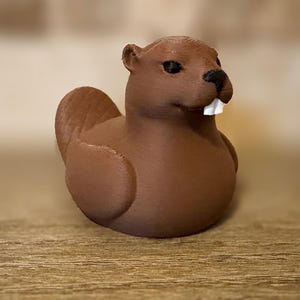 May include: A brown 3D-printed beaver figurine. The beaver has black eyes, a black nose, and two white front teeth. The figurine is sitting on a wooden surface.