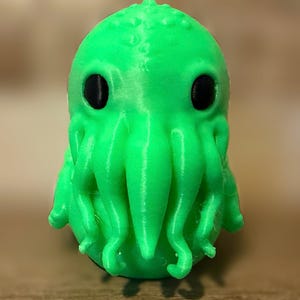 Peut inclure: Une figurine imprimée en 3D vert vif représentant une créature dotée d'une tête arrondie, de bosses texturées sur le dessus et de grands yeux noirs lisses. La figurine présente une série de tentacules pendants couvrant le bas du visage.