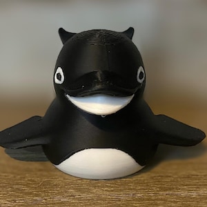 Puede incluir: Figura de orca impresa en 3D en blanco y negro. La orca tiene una boca sonriente, ojos pequeños y aletas en forma de aletas. La figura está sobre una superficie de madera.