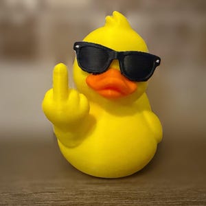 Puede incluir: Una figura de pato de goma de color amarillo brillante con gafas de sol negras. El pato tiene un pico naranja y está posicionado con un ala levantada, mostrando un gesto con el dedo corazón. La figura es lisa y tiene una textura impresa en 3D.