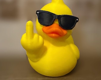 Op de afbeelding: Een felgeel rubberen eendje met een zwarte zonnebril. Het eendje heeft een oranje snavel en is gepositioneerd met één vleugel omhoog, waarbij een middelvingergebaar wordt gemaakt. Het figuurtje is glad en heeft een 3D-geprinte textuur.
