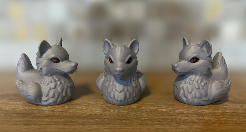 Three gray 3D printed duckies shaped like wolves with red eyes. The duckies are sitting on a wooden surface. Rubber Duck style.