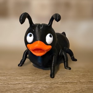 May include: A black 3D printed figurine shaped like an ant with a bright orange duck bill. The creature features two curved antennae on its head, large white eyes with black pupils, and six articulated legs extending from its rounded body.