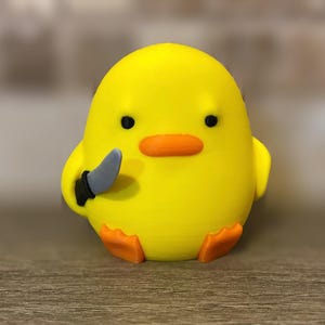 以下が含まれることがあります： オレンジ色のくちばしと足を持つ、黄色いひよこ型の置物。黒い柄の銀色のナイフを持っています。ひよこは黒い目を持ち、丸みを帯びた体です。ノベルティグッズです。
