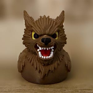 Puede incluir: Un pato de goma marrón con forma de hombre lobo, con orejas puntiagudas, ojos amarillos y una boca gruñendo que muestra dientes blancos y un interior rojo. La figura presenta detalles de pelaje texturizado en la cabeza y el pecho.