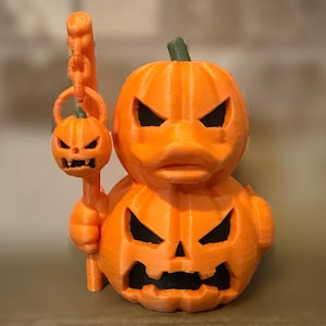 May include: A bright orange 3D printed duck figure shaped like a stack of two carved jack-o'-lanterns. The duck holds a tall staff topped with a smaller, matching jack-o'-lantern head. Both pumpkins feature sharp, triangular eyes and jagged mouths.