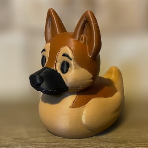 Op de afbeelding: Een 3D-geprinte badeend in de vorm van een Duitse herdershond. De eend heeft een bruin lichaam met bruine en zwarte details op de kop en oren. De eend heeft een zwarte neus en ogen.