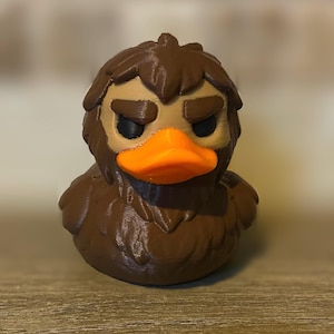 May include: A 3D-printed duck figurine with a brown body and head, featuring a tan face and an orange beak. The duck has a stern expression, with furrowed brows and a slightly grumpy look. The figurine is a unique decorative item.