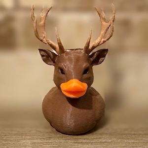 Könnte beinhalten: Eine skurrile braune Hirsch-Ente-Hybridfigur mit großem Geweih und einem orangefarbenen Schnabel. Die Figur ist 3D-gedruckt und hat eine strukturierte Oberfläche. Die Hirsch-Ente ist ein einzigartiger Dekorationsartikel.