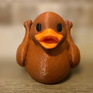 May include: A brown duck-shaped figurine with a bright orange beak and black eyes. The figure has a rounded body and two raised wing-like appendages on either side of its head, giving it a distinct appearance with a textured surface finish.