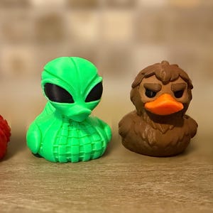 May include: Four novelty rubber duck toys in a row. The ducks are red, green, brown, and green. Each duck has a unique design, including a bird, an alien, a duck with a frown, and a snake-like duck.