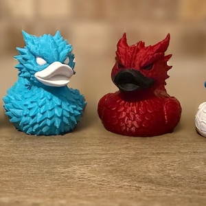 Puede incluir: Cuatro patos de goma con temática de dragones en fila. Los patos son en tonos grises, azules, rojos y blancos. Cada pato tiene un diseño único de dragón, con detalles y expresiones. Se exhiben sobre una superficie de madera.