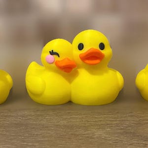 Three 3D printed couples Duck of a girlfriend and a boyfriend all yellows ducks connected with orange beaks and black eyes. Girlfriend duck has eyelashes and pink Rosie cheek. Rubber Duck style perfect for jeep ducking. Facing front.
